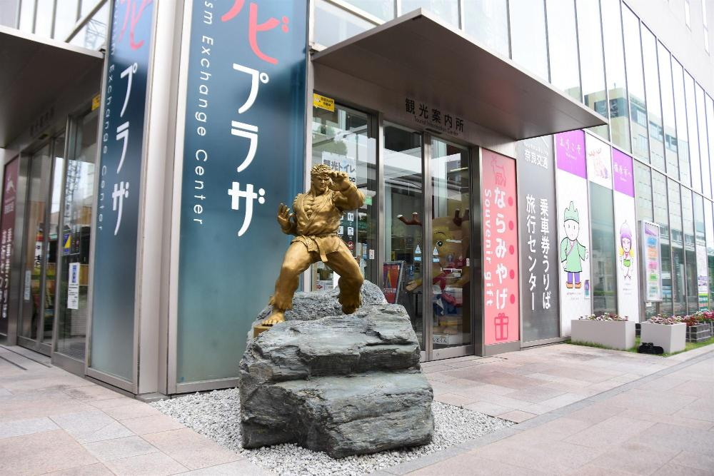 Estátua de bronze dinâmica de Ryu em pose de ação, colocada em frente a um centro de troca turística com escrita em japonês.